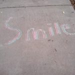 concrete sidewalk with blue, pink, and Yellow chalk drawing the word "smile".