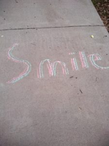 concrete sidewalk with blue, pink, and Yellow chalk drawing the word "smile".