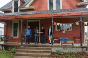 4 masked and social distanced people standing on porch of 2 story orange house, receiving a donation to help feed the neighbor to neighbor food for families program.