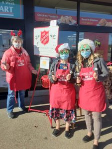 3 ladies ringing the Salvation Army bell in front of a grocery store. the 2 teenaged girls are wearing fun, sparkly glasses with santa hats. The "mature" woman is wearing a headband with reindeer antlers.