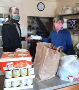 Tall man with beard and mask handing woman wearing a pink mask a check. The check was money raised during a local Frisbee tournament. Also shown are food items that were also donated along with the money.