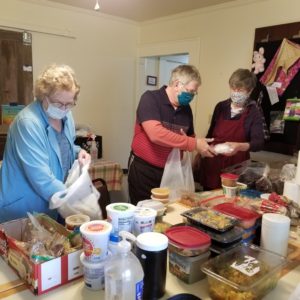 2 females and 1 male, all wearing masks and gloves packing free meals into plastic bags for disbursement to the community.